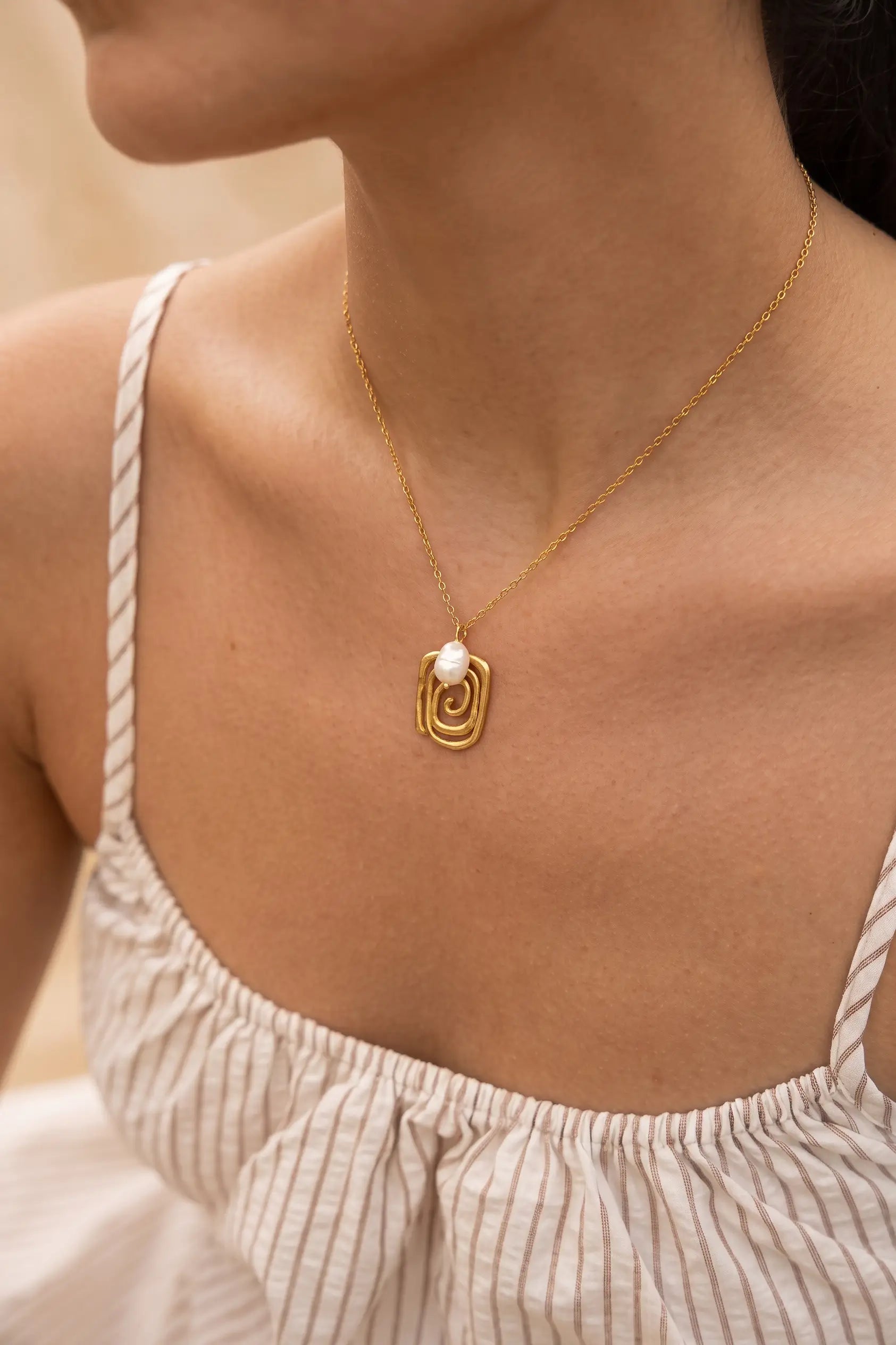 A close-up of a woman wearing a delicate gold chain necklace with a small gold spiral pendant and a white pearl accent, paired with a cream-colored striped top.