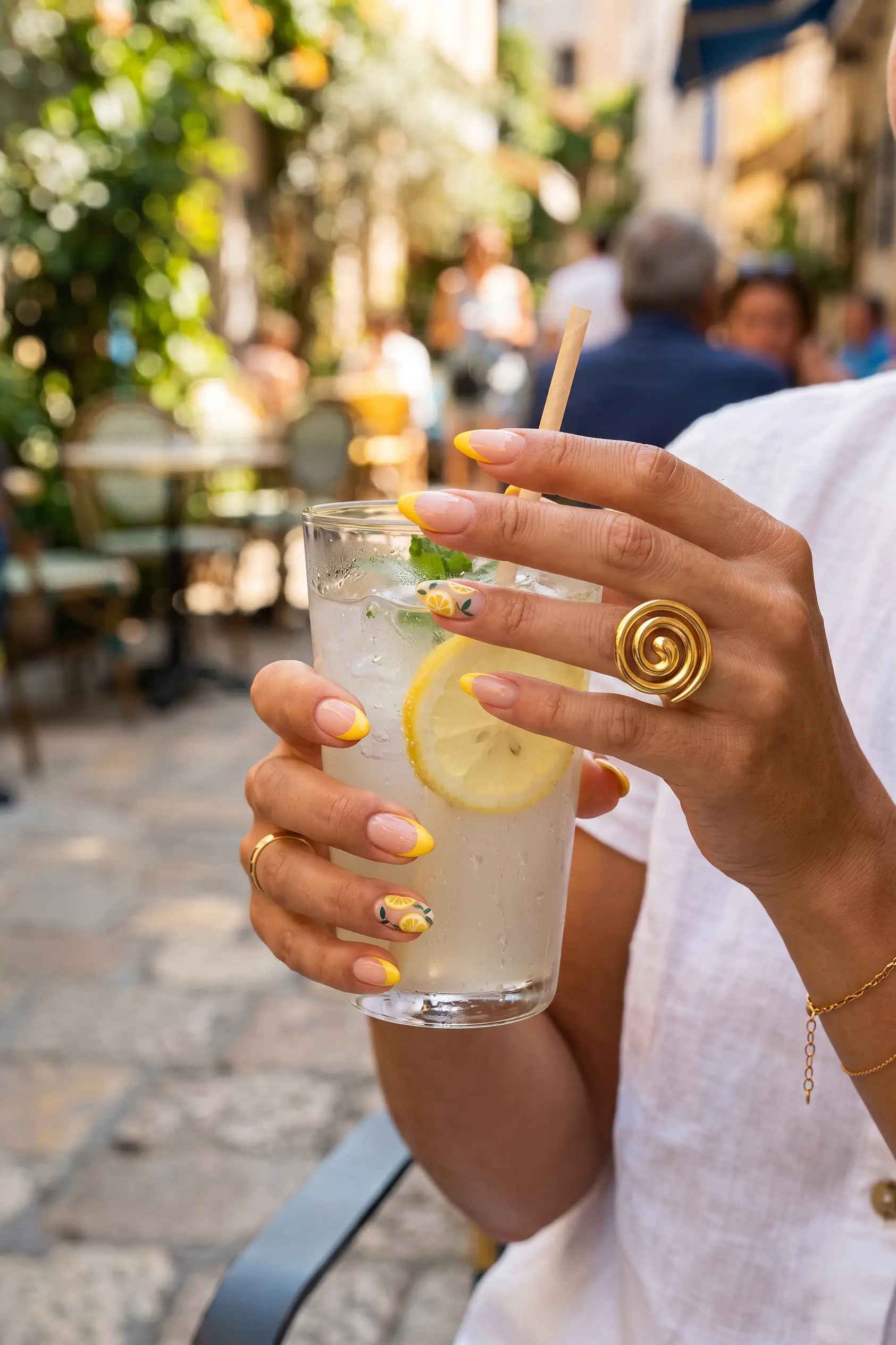 A close-up of a person holding a glass of lemonade with a straw in an outdoor cafe setting. The person is wearing a white top and gold jewelry, including a large spiral ring on one finger.