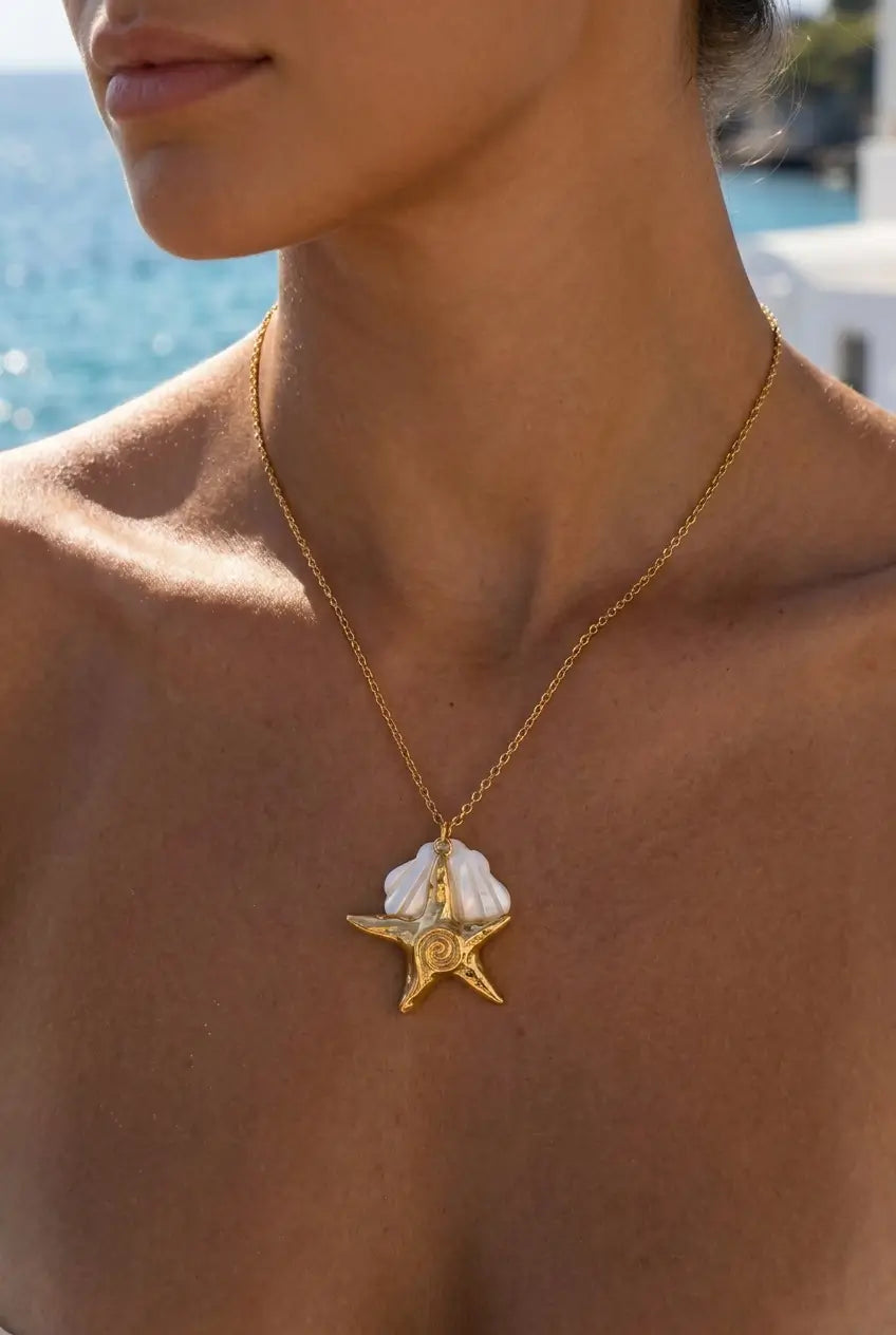 A gold necklace with a starfish and shell pendant worn by a model outdoors near the ocean.