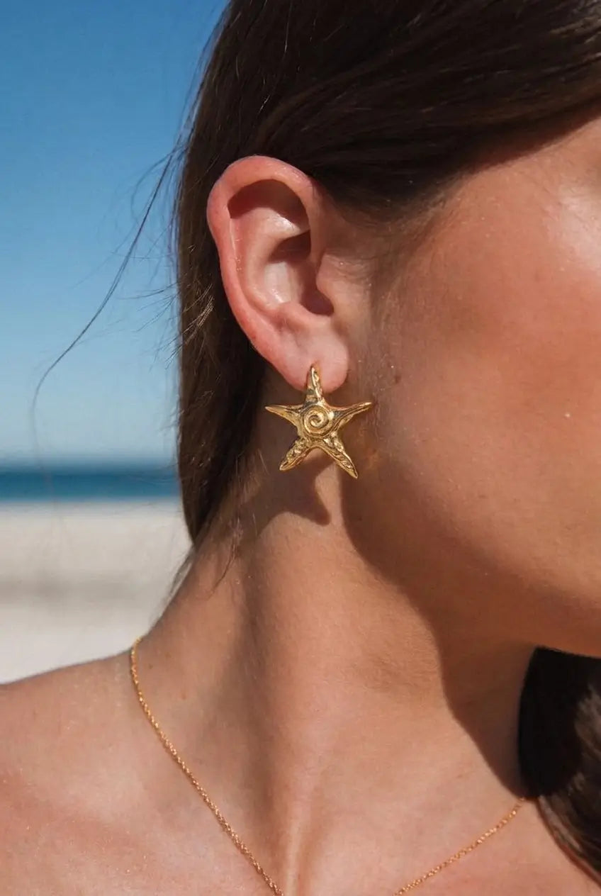 A close-up of a woman's ear wearing a gold starfish-shaped stud earring, with a beach and blue sky blurred in the background.
