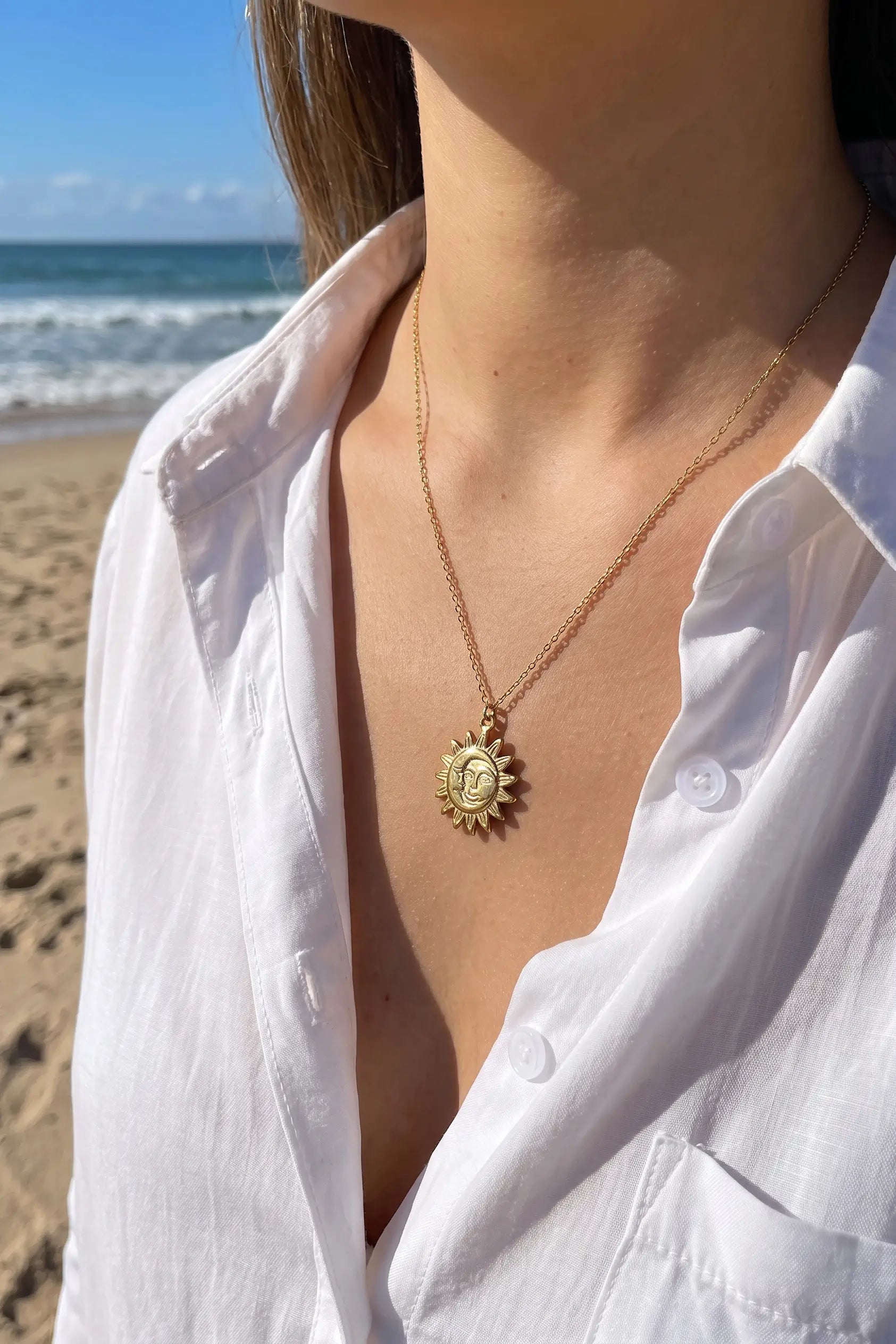 A close-up lifestyle shot of a woman wearing a gold pendant necklace with a sunburst design and a medallion pendant, set against a beach background with ocean and sand visible.
