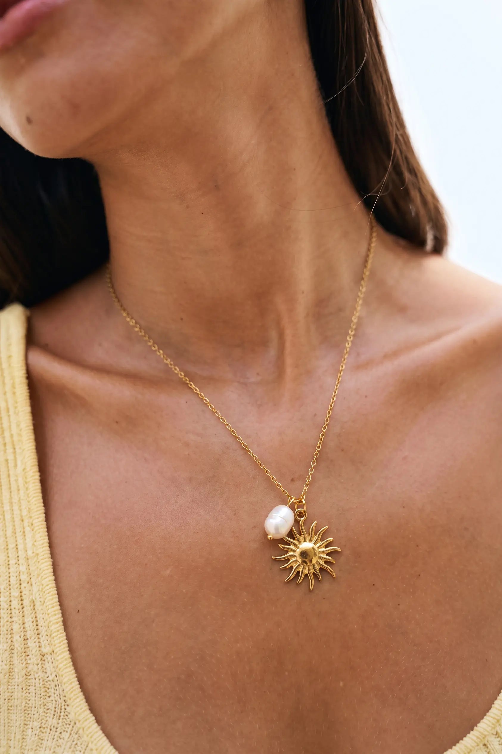 A close-up of a woman wearing a delicate gold chain necklace with a gold sunburst pendant and a white baroque pearl, paired with a yellow top.