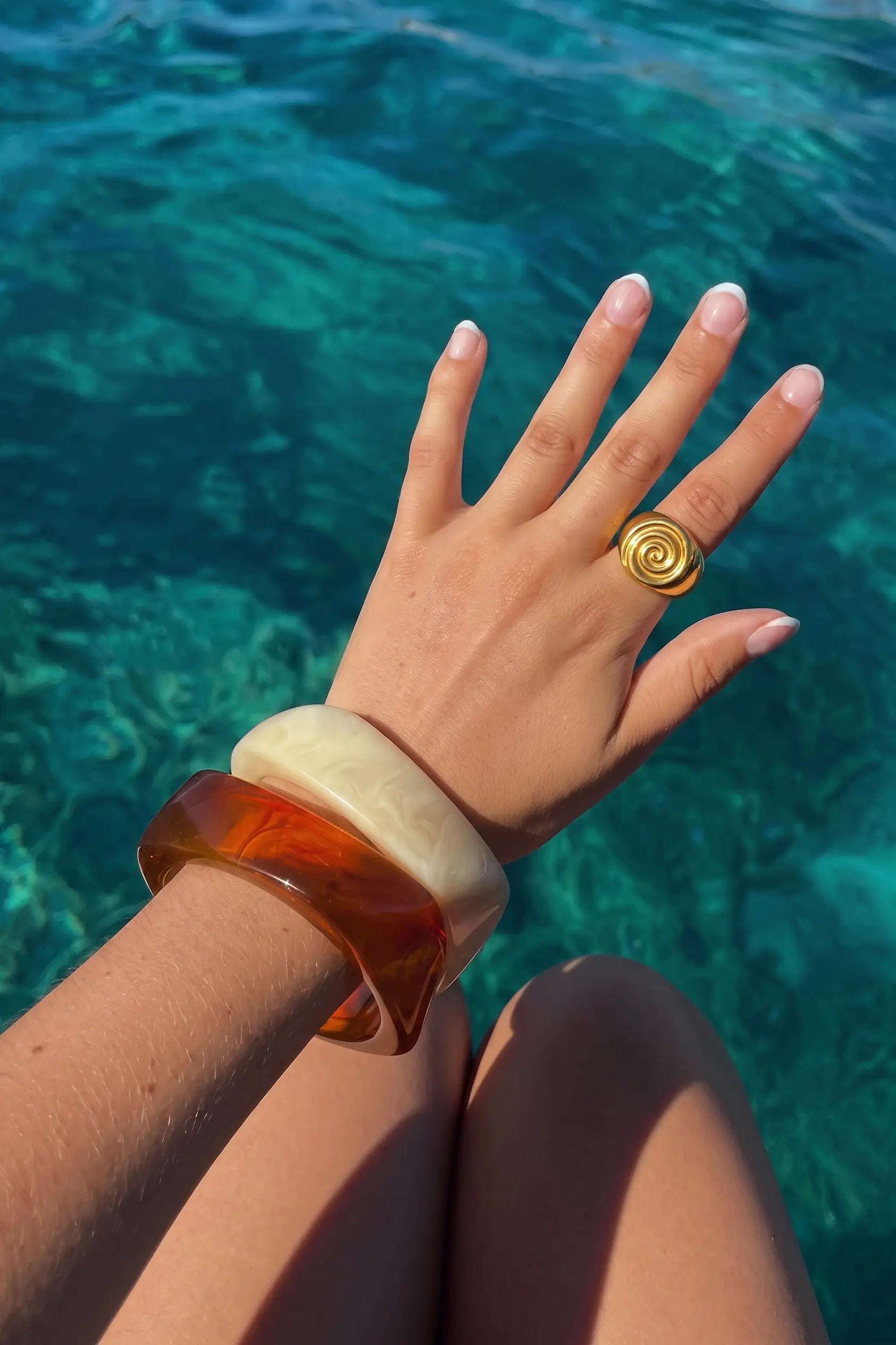 A close-up of a person's hand resting over clear turquoise water, wearing a chunky amber-brown translucent bangle and a gold spiral ring.