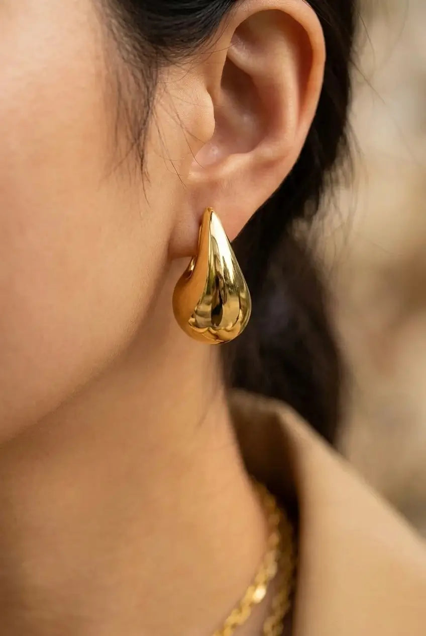 A close-up of a person's ear wearing a shiny gold teardrop-shaped hoop earring, with part of a beige top and a gold chain necklace visible against a softly blurred background.
