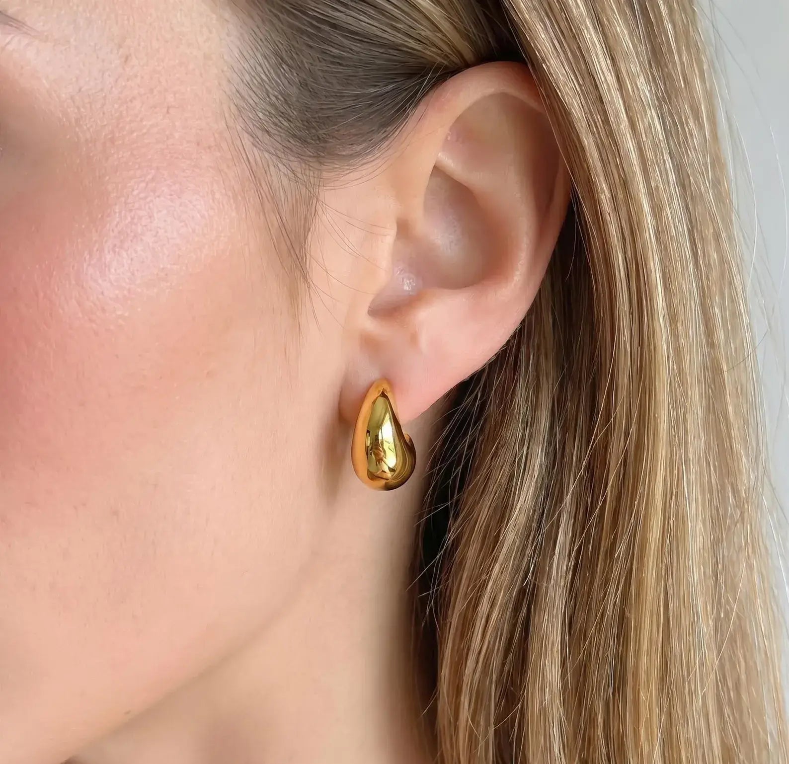 A close-up of a woman's ear wearing a shiny gold teardrop-shaped stud earring.