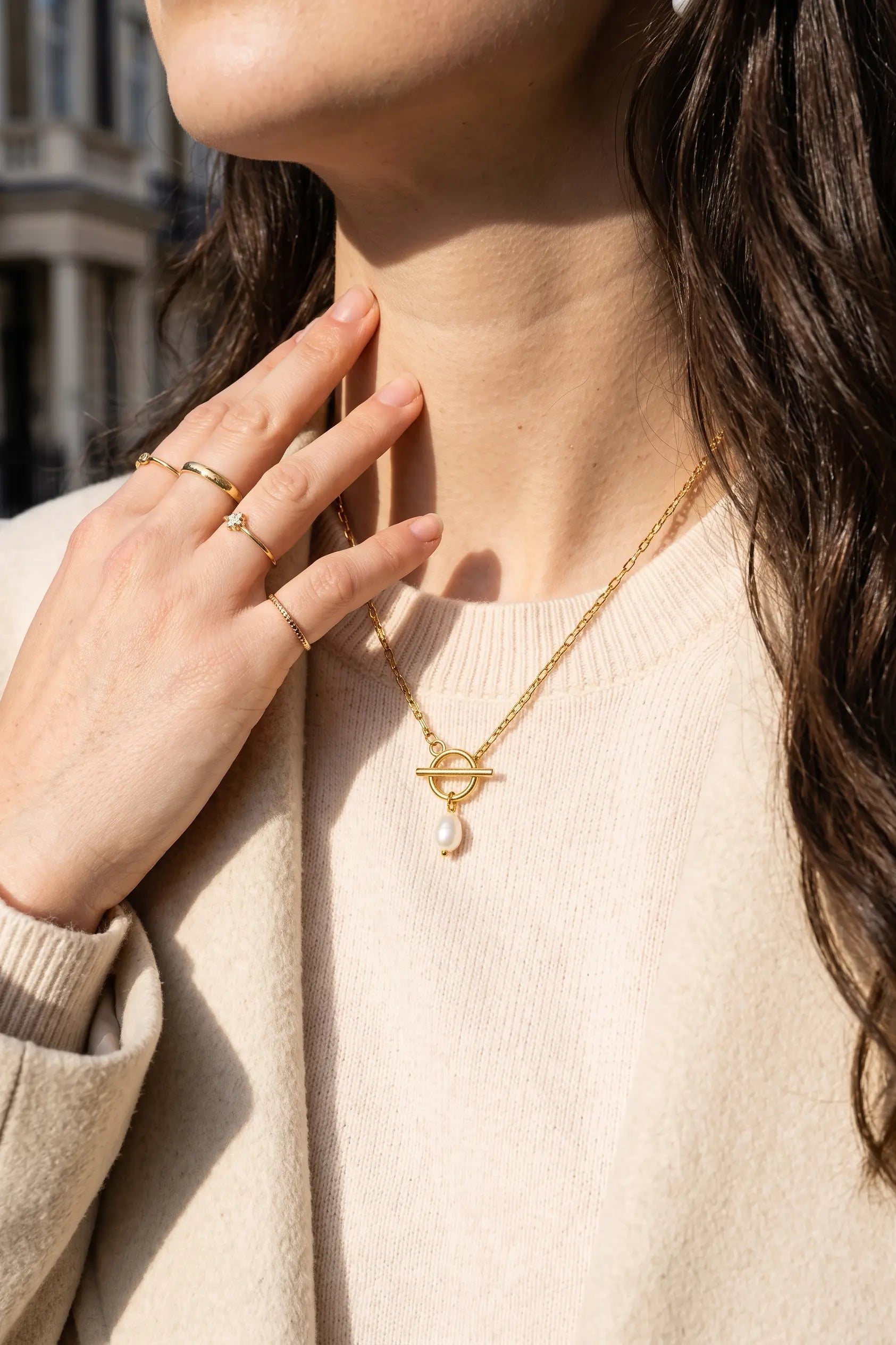 A close-up lifestyle shot of a woman wearing a delicate gold necklace with a geometric eye-like pendant and a small pearl drop, styled with gold rings on her fingers.