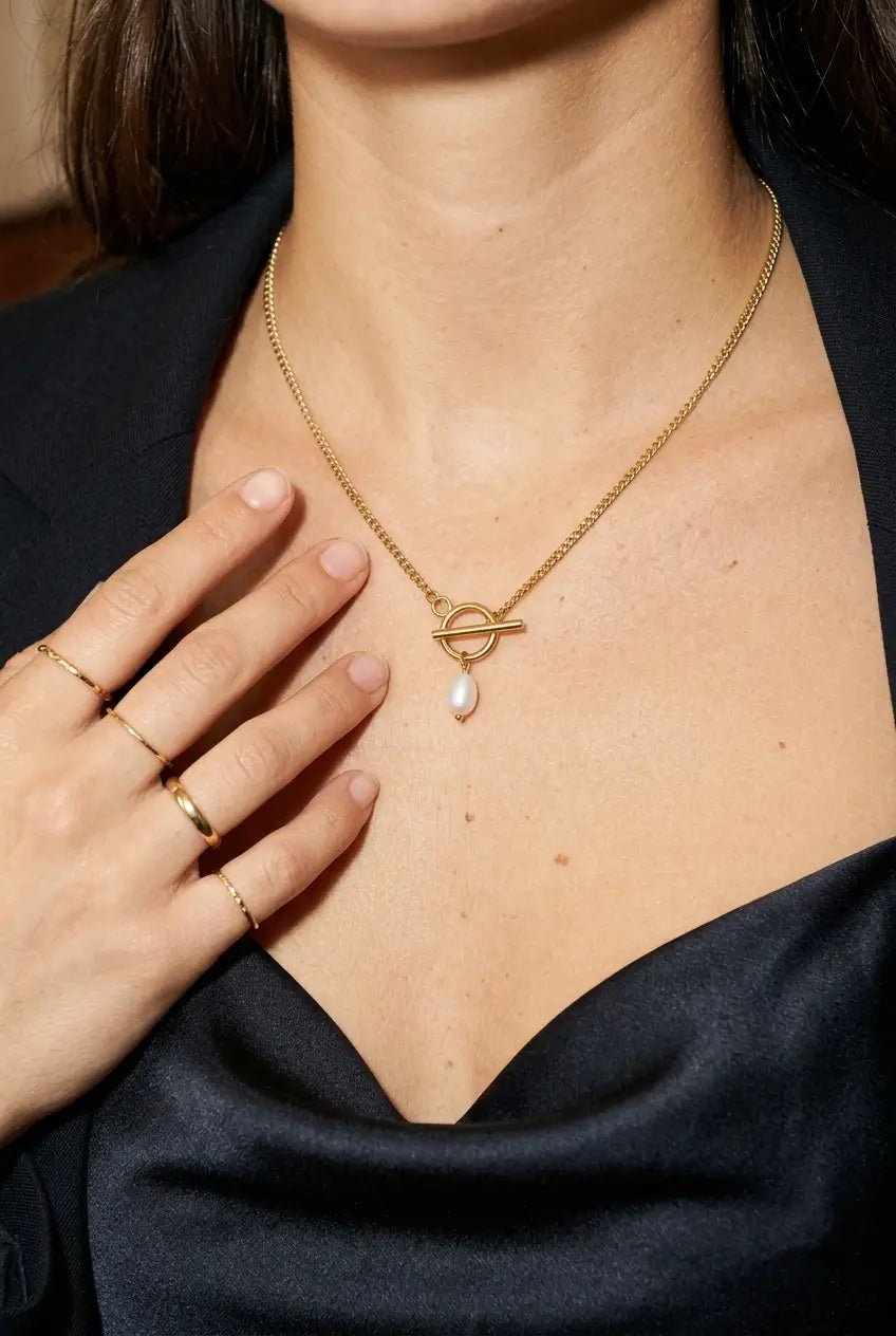 A close-up of a model wearing a delicate gold chain necklace with a circular toggle pendant and a small teardrop pearl drop. The model is wearing a black satin top and has gold rings on her fingers.