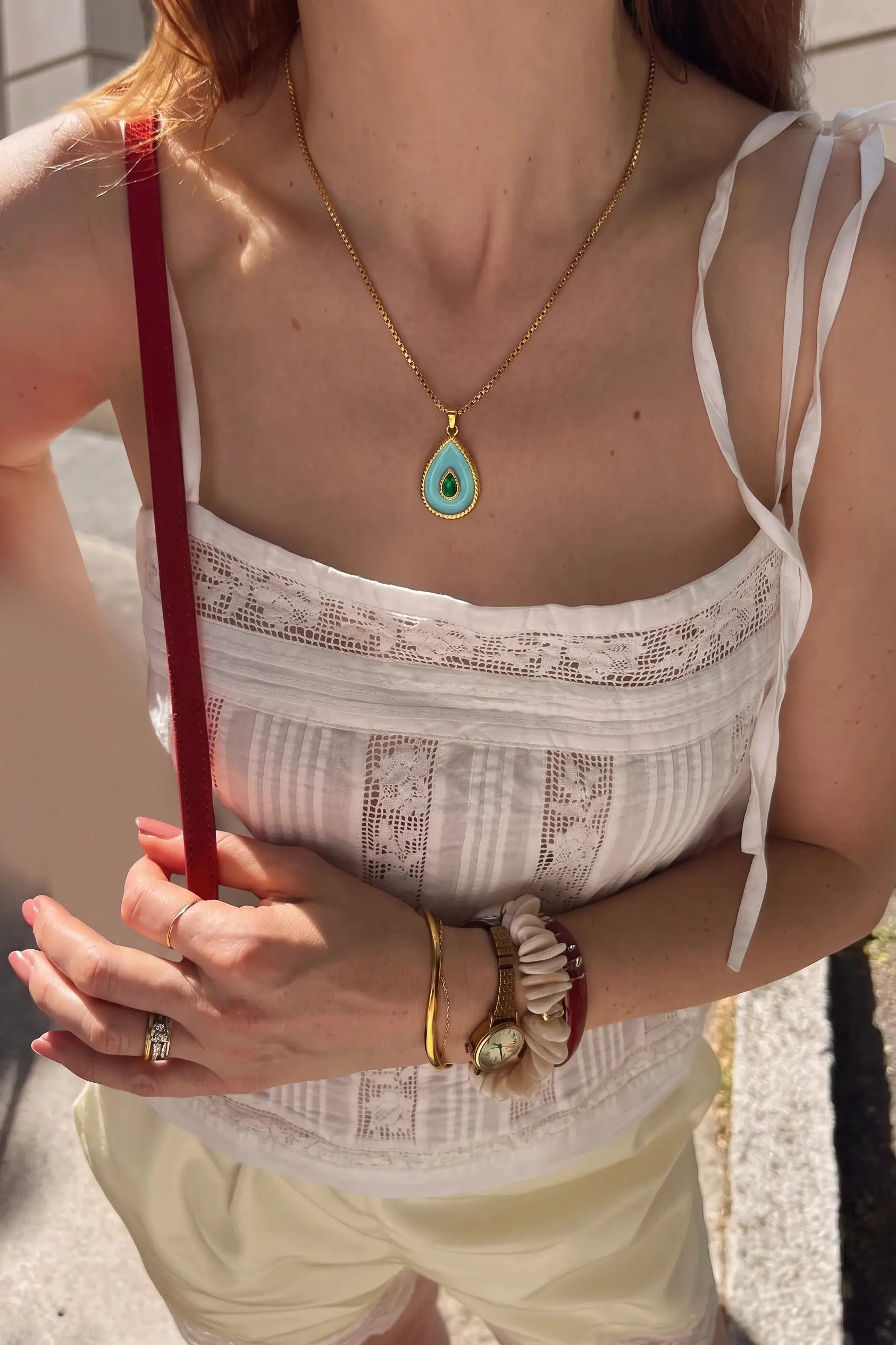 A close-up lifestyle shot of a woman wearing a gold teardrop pendant necklace with a turquoise-colored stone, styled with a white lace camisole top and cream shorts.