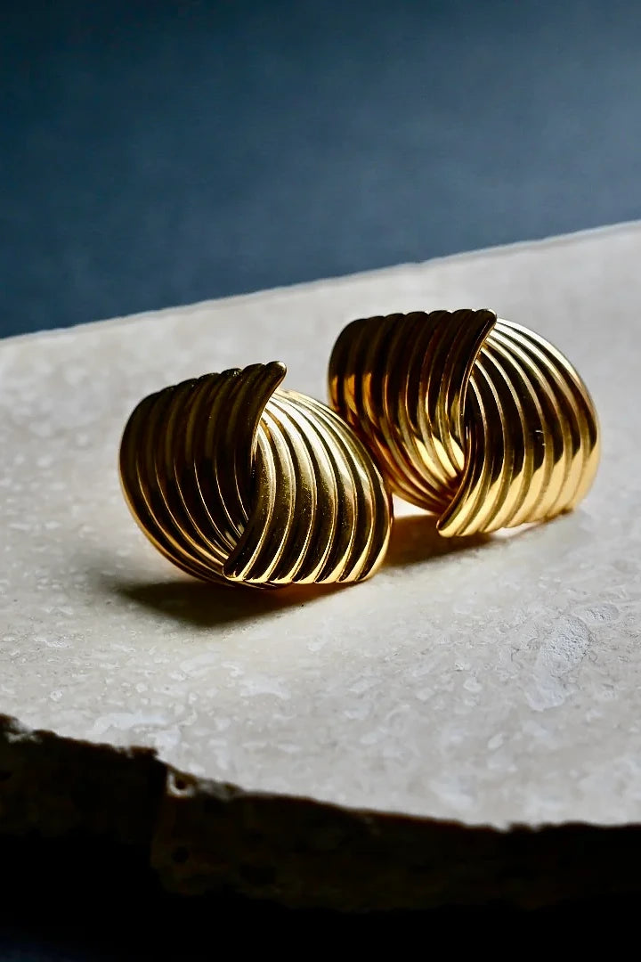 A pair of gold-toned, ribbed spiral hoop earrings resting on a textured white stone slab against a dark blue background.