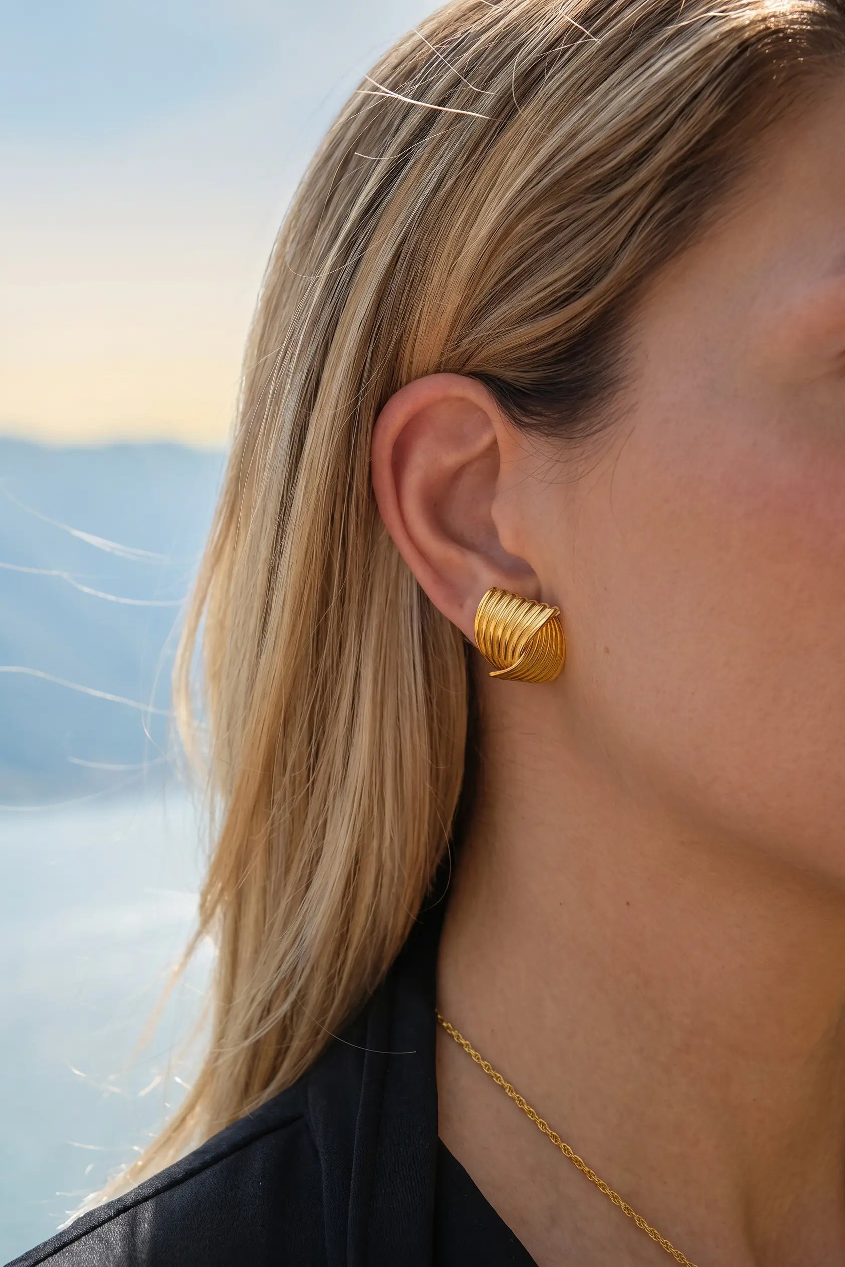 A close-up side profile of a woman with blonde hair wearing a textured gold hoop earring and a delicate gold chain necklace, set against a soft-focus outdoor background.