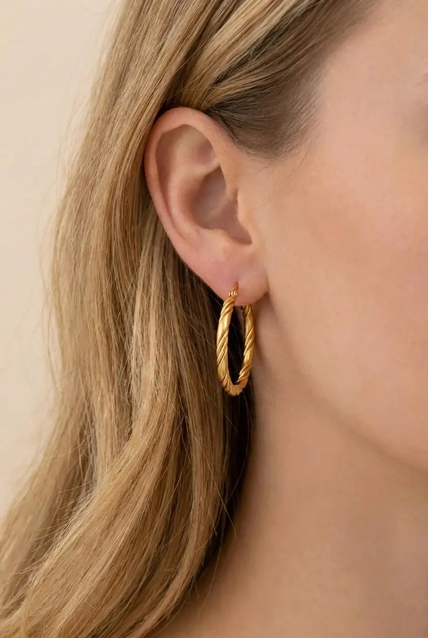A close-up of a model's ear wearing a pair of textured gold hoop earrings against a soft beige background.