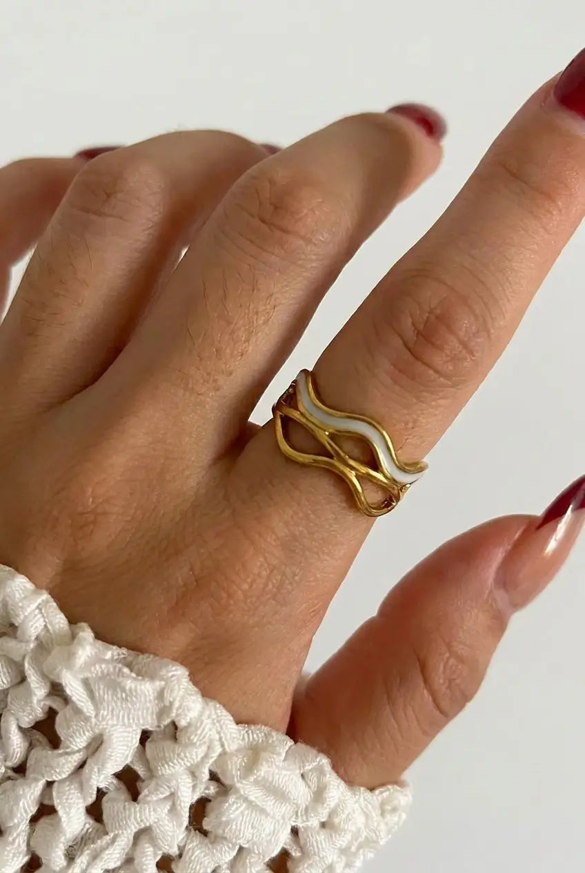 A close-up of a hand wearing a gold wavy open ring, with dark red manicured nails and a white textured knit cuff visible at the wrist, against a plain light background.