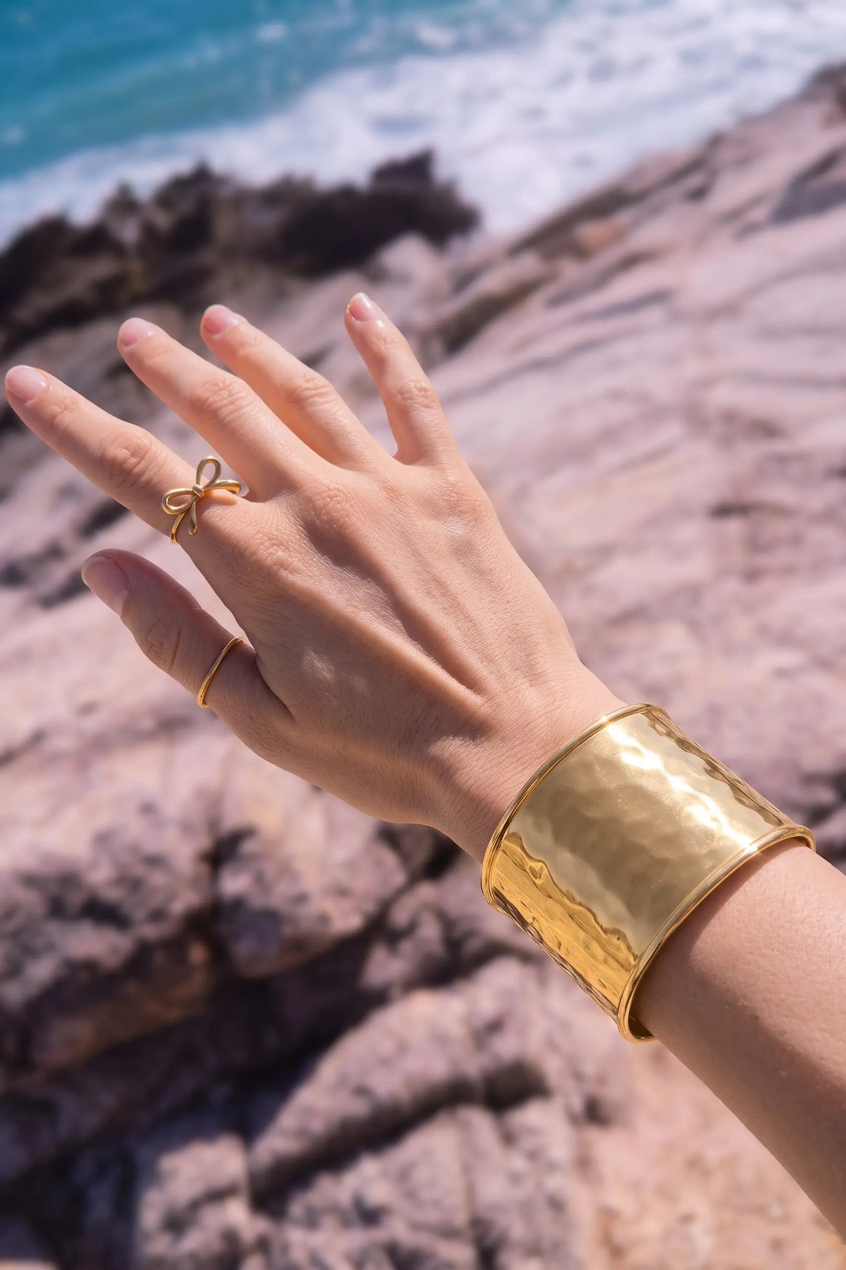 A close-up of a person's hand and wrist wearing a wide, hammered gold cuff bracelet and a small gold bow ring, set against a blurred rocky beach and ocean background.