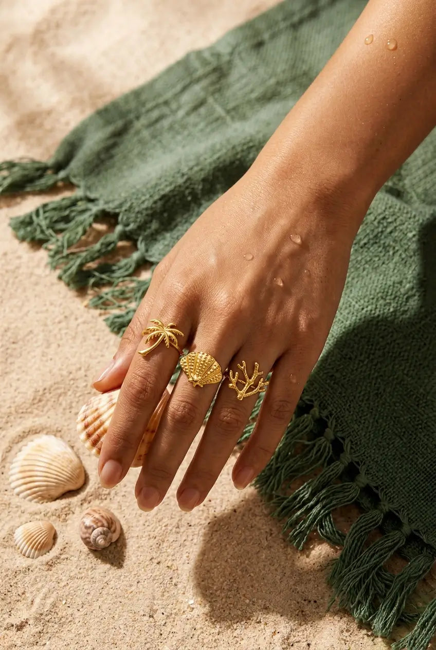 A close-up of a person's hand resting on sandy beach ground next to a green fringed towel, wearing three gold rings: a palm tree ring, a textured shell ring, and a coral ring.