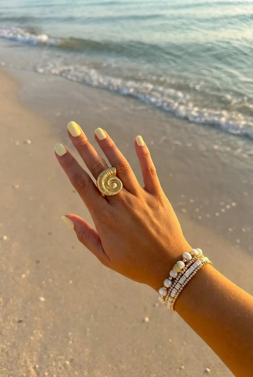 A close-up of a person's hand on a sandy beach, wearing a large gold spiral shell ring and pearl bracelets.