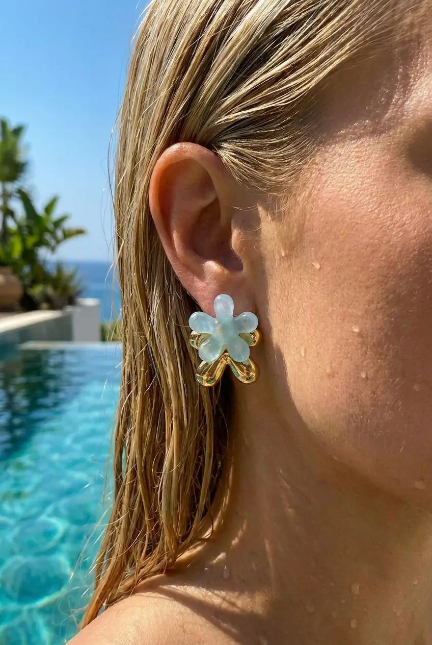 A close-up lifestyle shot of a woman's ear wearing a gold flower-shaped stud earring with a light blue cabochon center, set against a sunny poolside background.