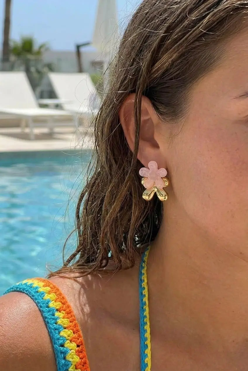 A close-up lifestyle shot of a woman by a swimming pool wearing a pink flower-shaped earring with gold accents.