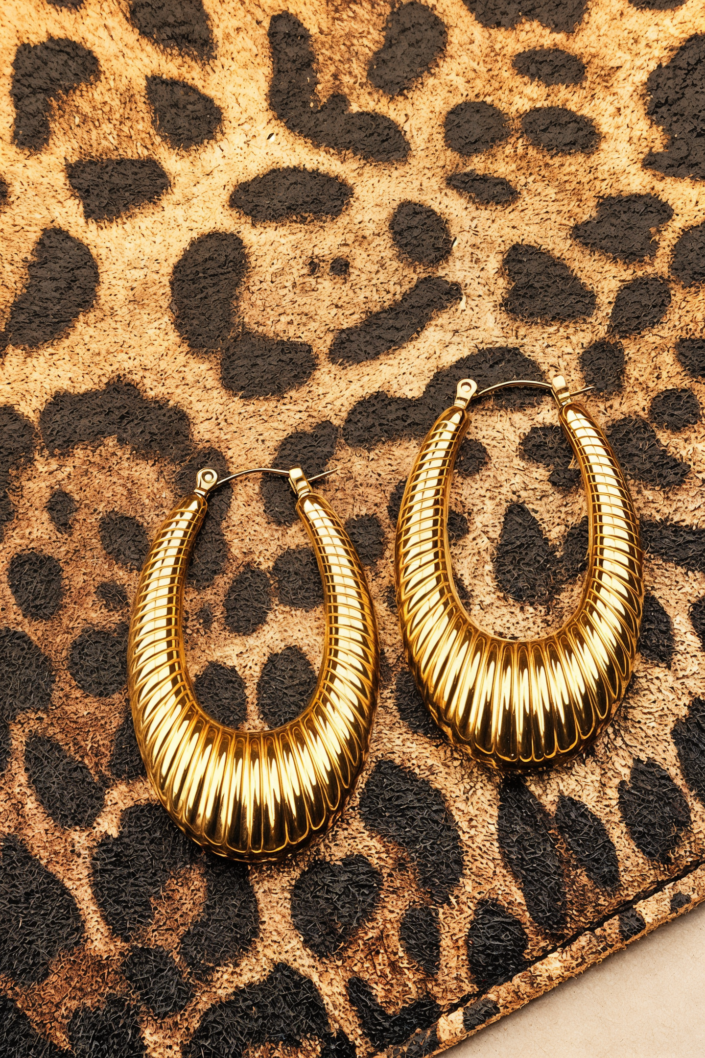 A pair of large gold hoop earrings with a ribbed, textured finish, displayed on a leopard print surface.
