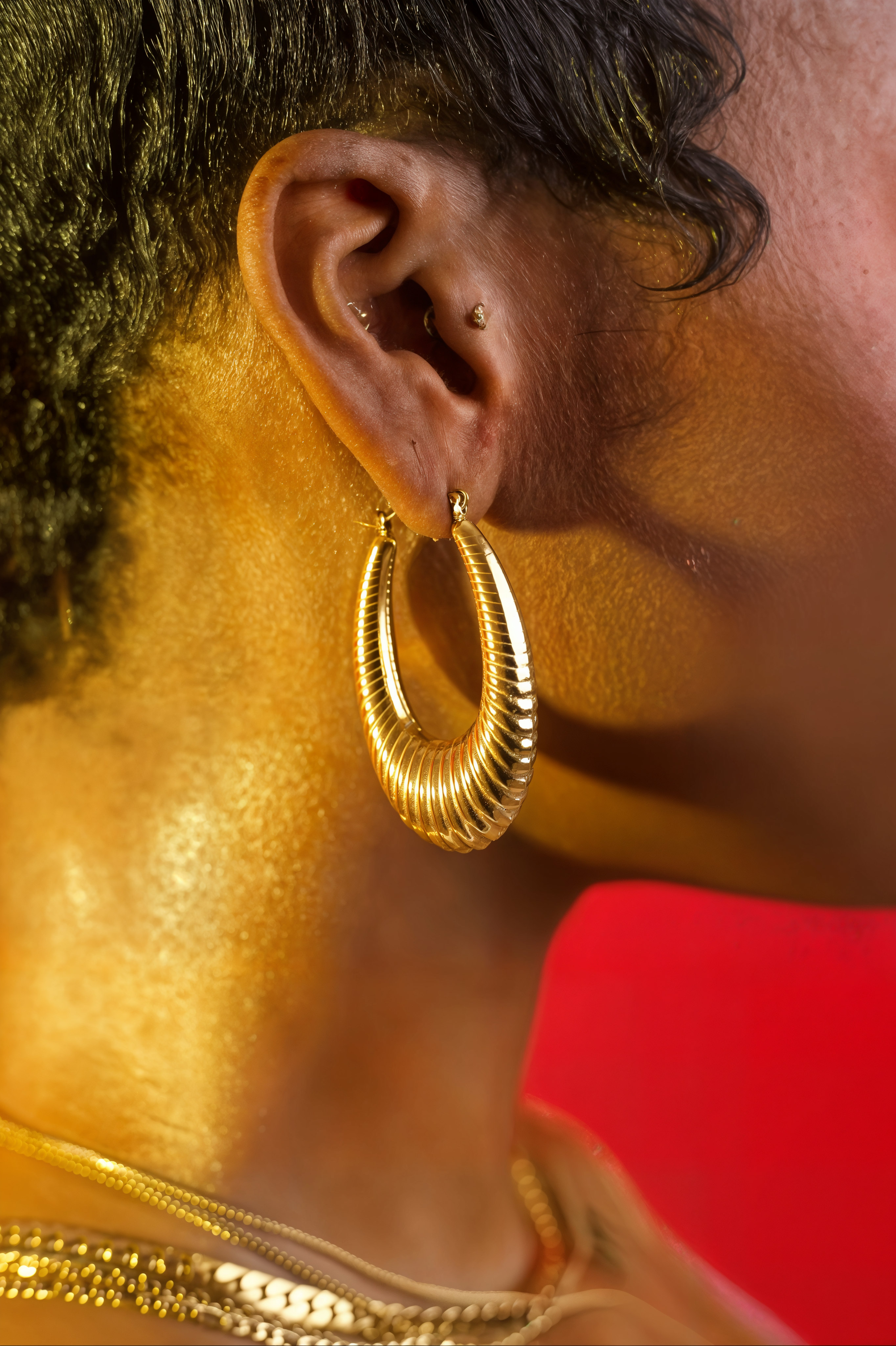 A close-up side profile of a person wearing a large textured gold hoop earring, with gold necklaces visible and a bright red background.