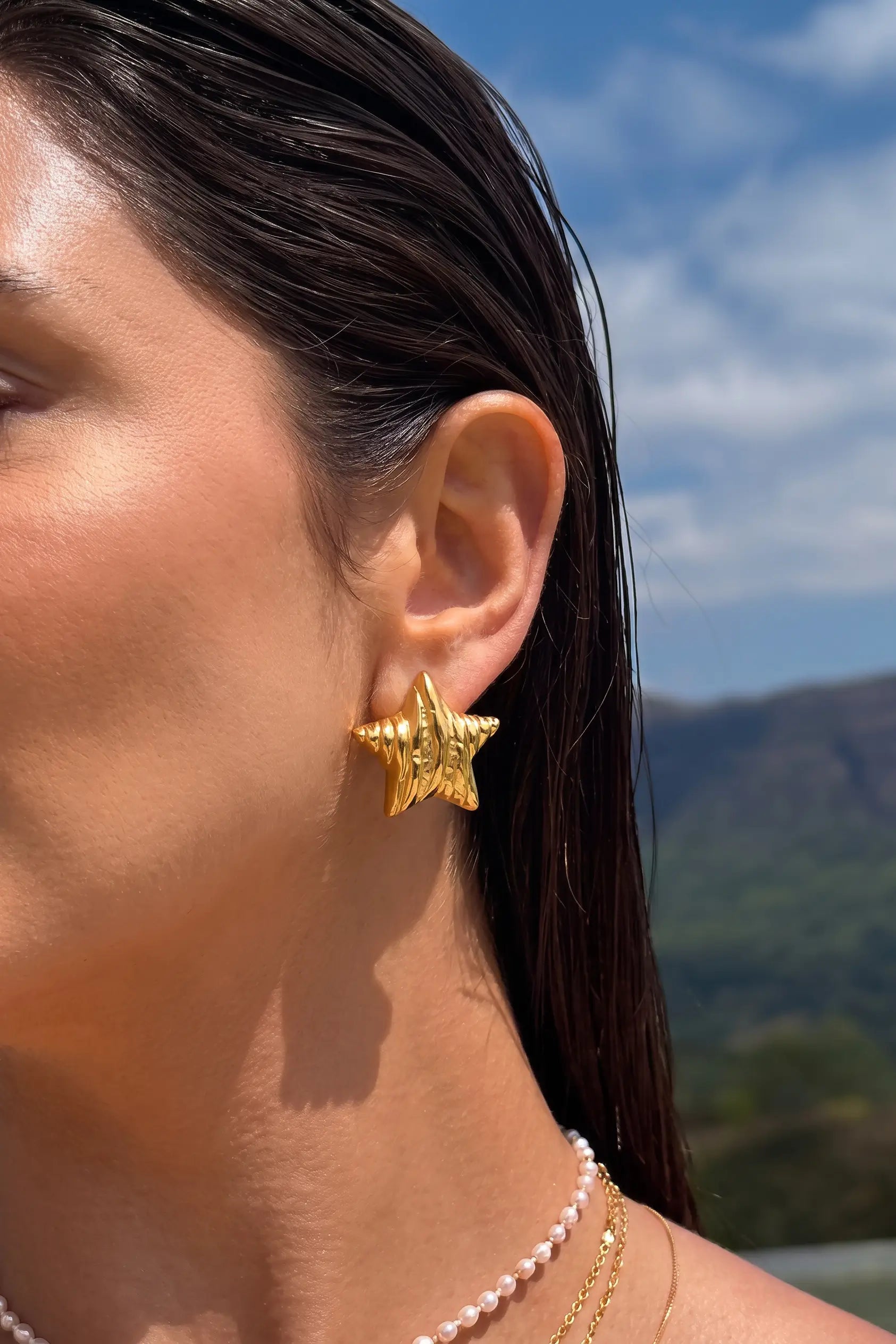 A close-up side profile of a woman wearing a textured gold star-shaped stud earring, with a pearl necklace and delicate gold chain visible, set against an outdoor background of blue sky and green hills.
