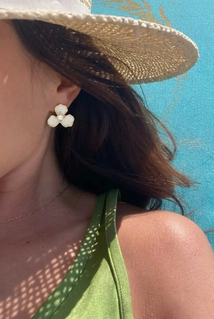 A close-up lifestyle shot of a woman wearing a white three-petal flower stud earring, a straw hat, and a green top against a bright blue background.