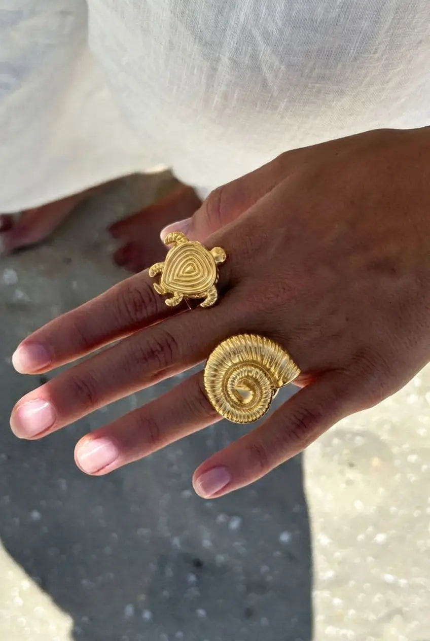 A close-up of a hand wearing two gold rings: a small turtle ring on the middle finger and a larger spiral shell ring on the ring finger, with a white garment and outdoor ground visible in the background.