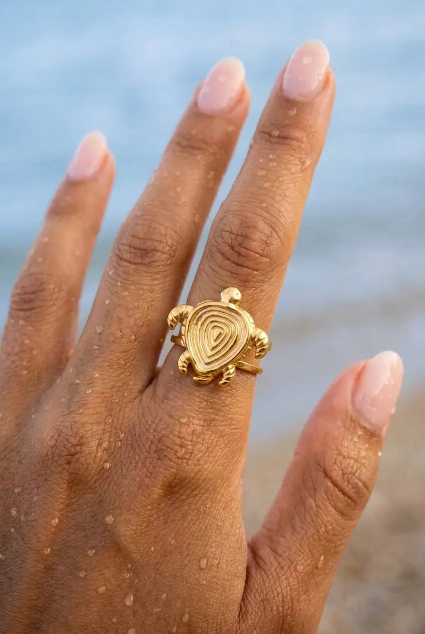 A gold turtle ring worn on a person's ring finger, shown close-up with a blurred beach and water background.