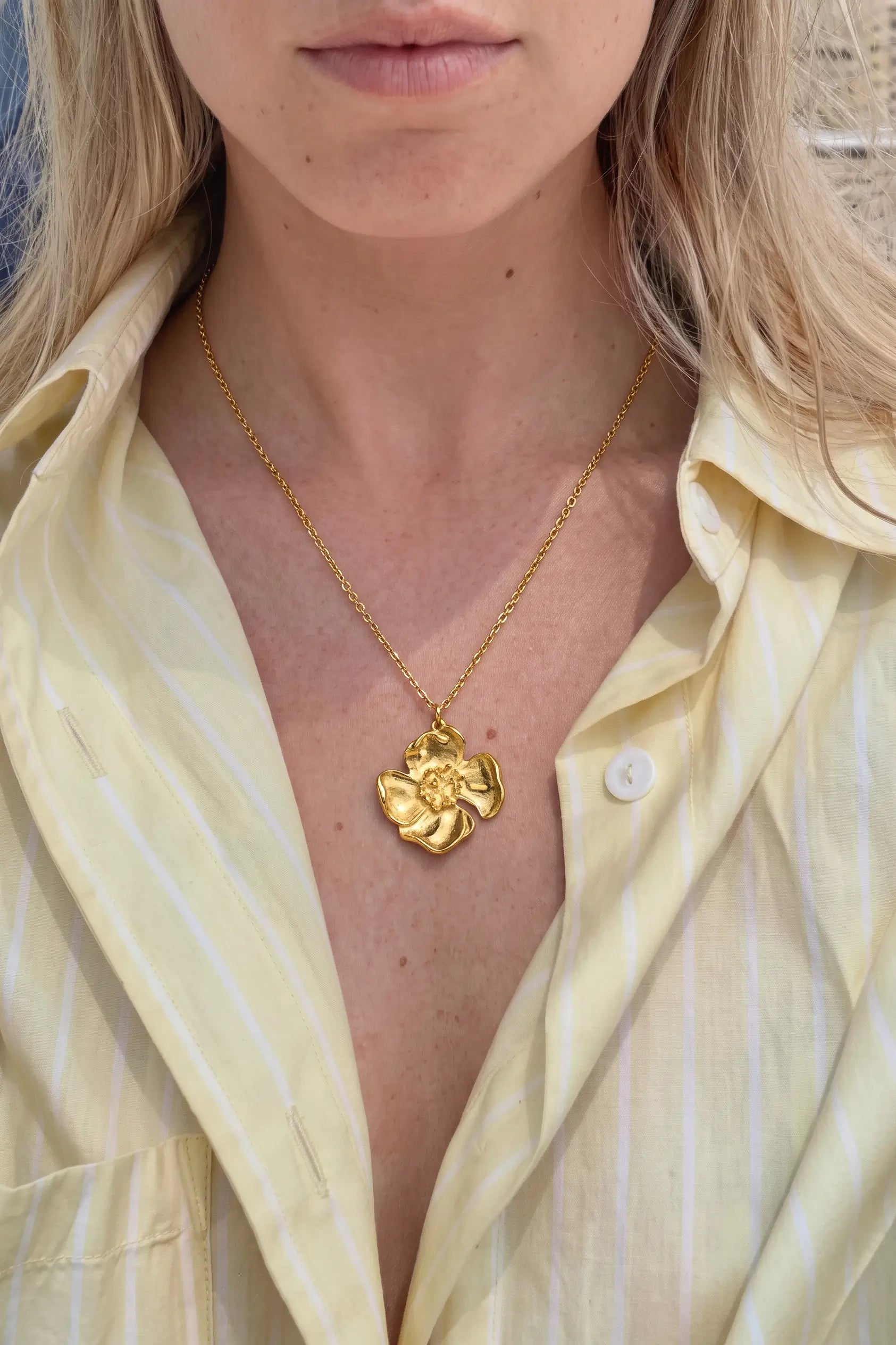 A close-up of a model wearing a gold flower pendant necklace on a delicate gold chain, paired with a pale yellow striped button-down shirt.