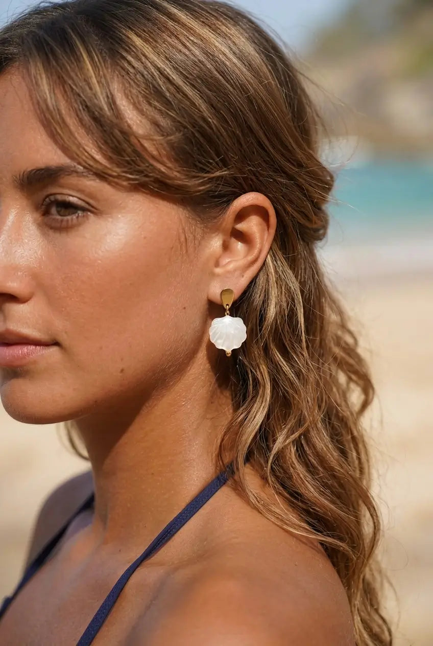 A woman with wavy light brown hair is shown in profile at a beach, wearing a single drop earring with a white flower-shaped charm and a gold top post.