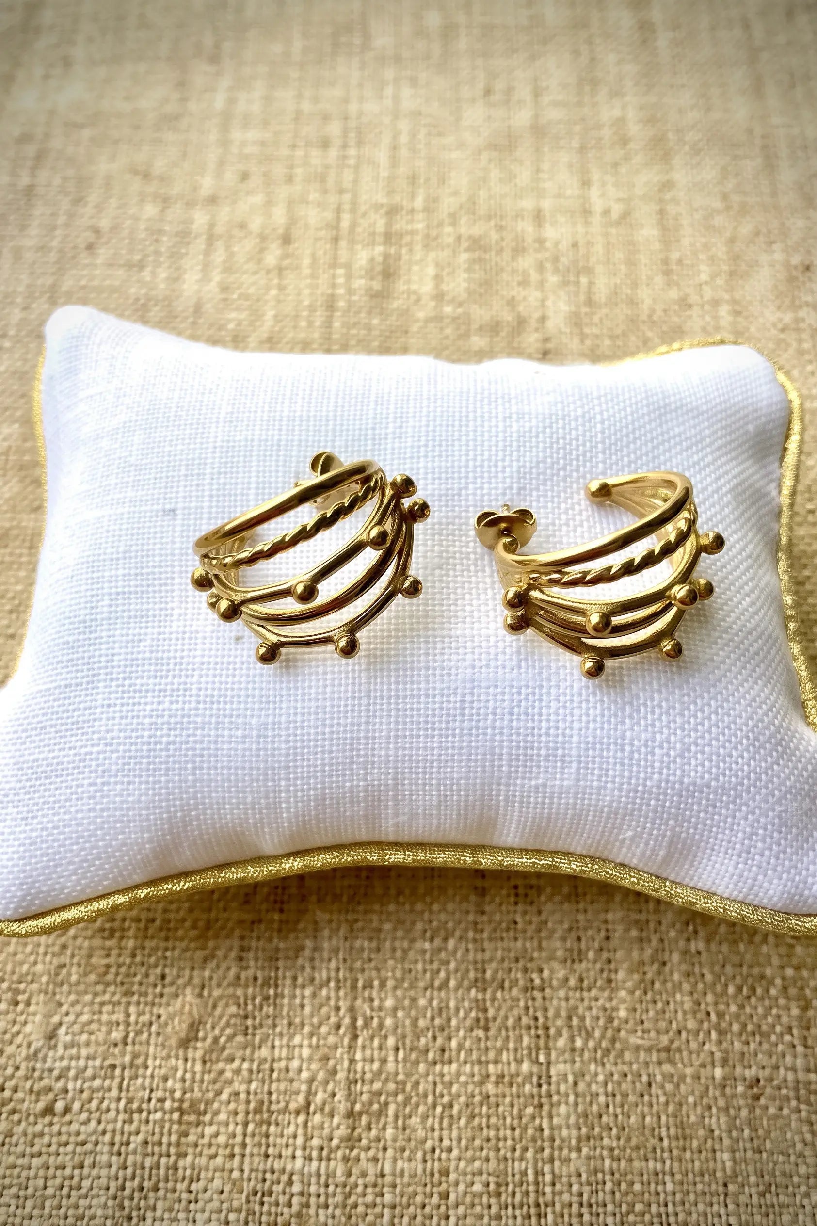 A pair of gold-toned multi-band hoop earrings displayed on a white fabric jewelry pillow with gold trim, set against a textured beige burlap background.